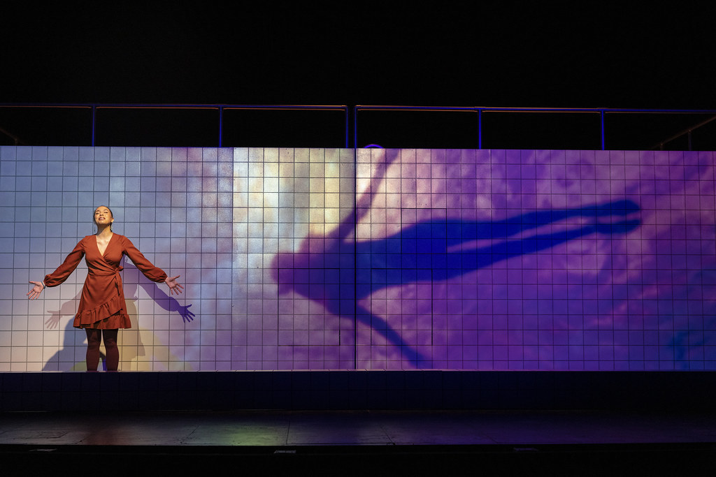 A woman stands, arms out, eyes closed; a projection of a female figure appears on the wall behind her in a similar pose.