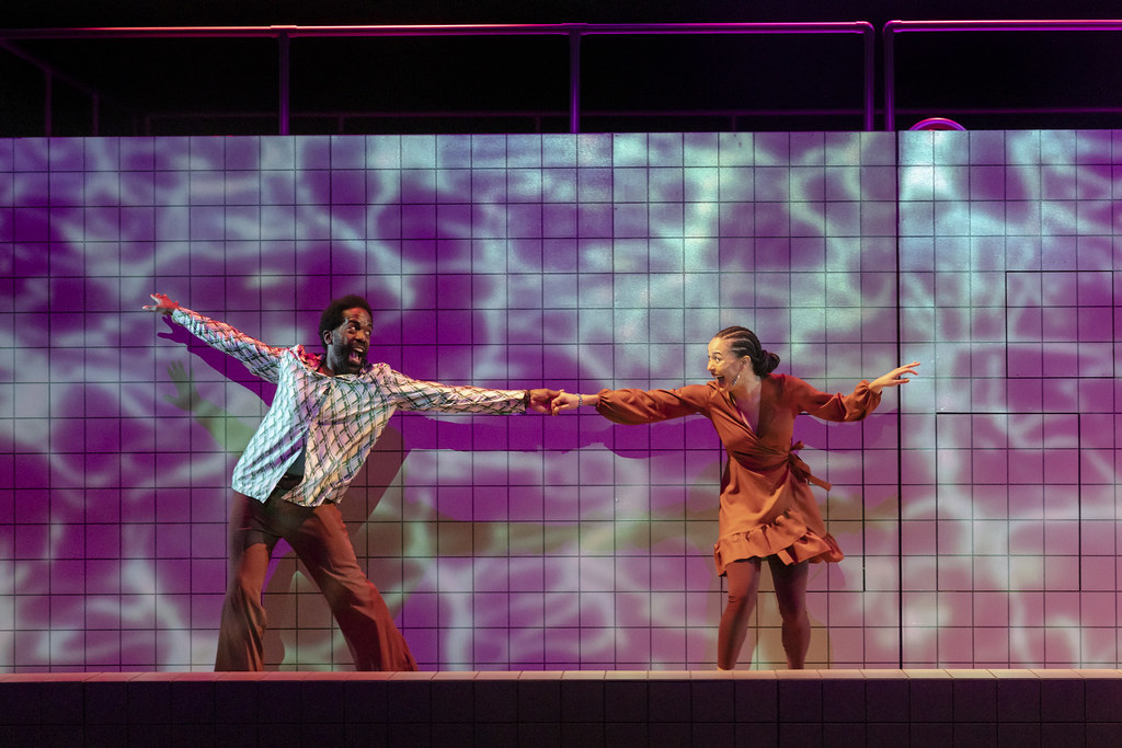 A man and a young woman split center stage; they dance, holding hands with one arm out each.