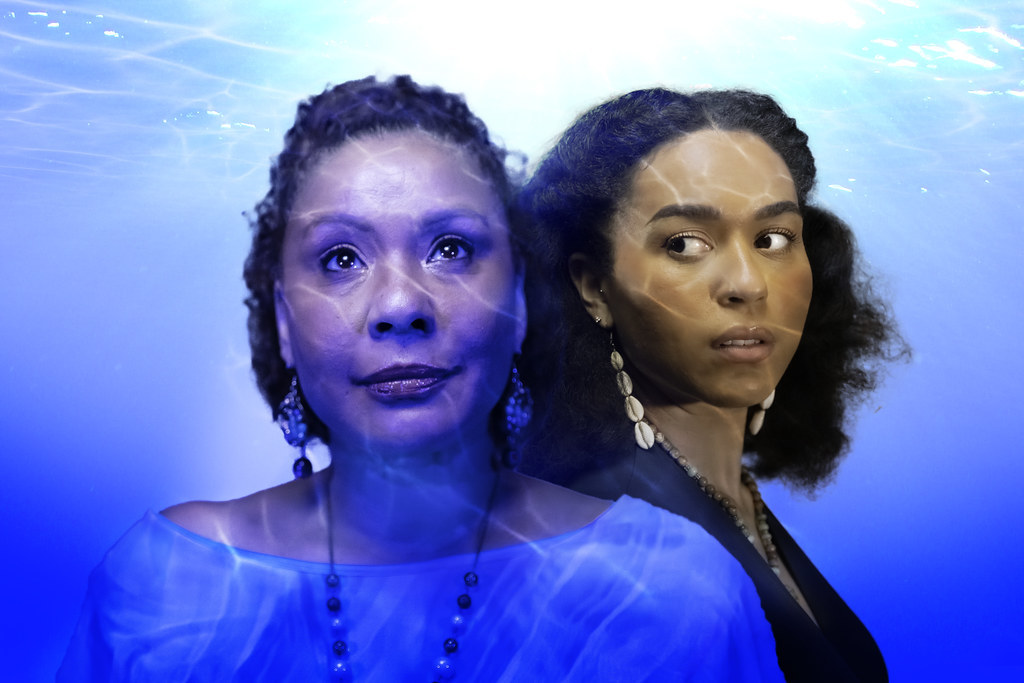 Two women stand submerged by water. The center looks up and off, while the other is behind her, looking over her shoulder.