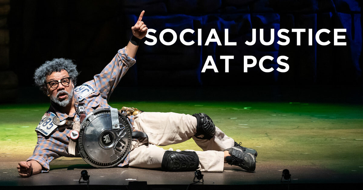 A man lies on his right side on the stage, his left arm raised with one finger pointed skyward.