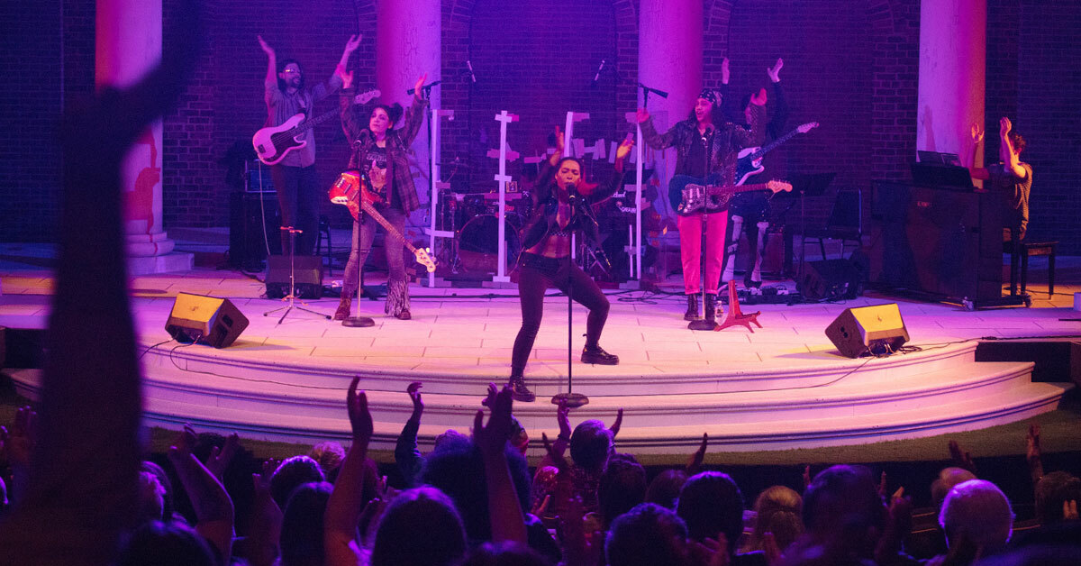 A rock band plays on stage as a crowd in the foreground watches, their hands in the air.