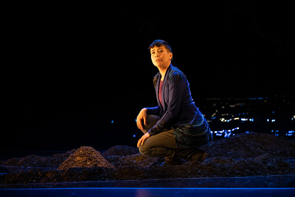 A woman kneels in a pool of light next to a small pile of rubble, her head turned toward the audience.