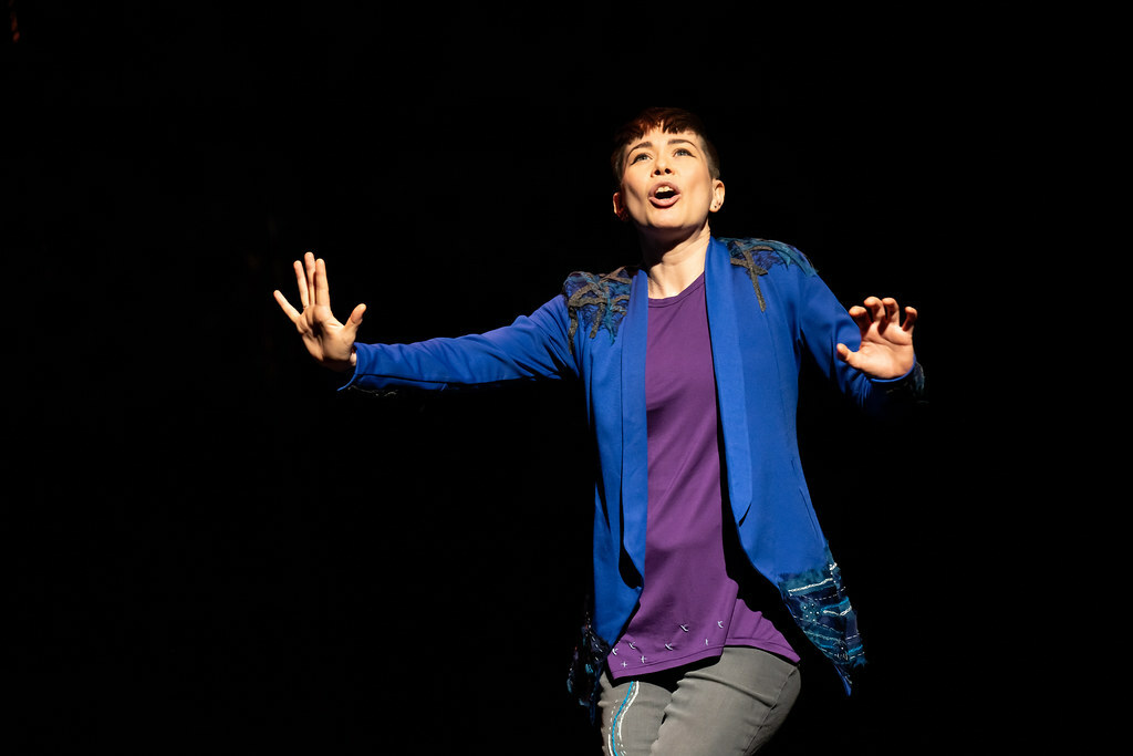 A woman stands against a dark background, speaking, her arms outstretched.