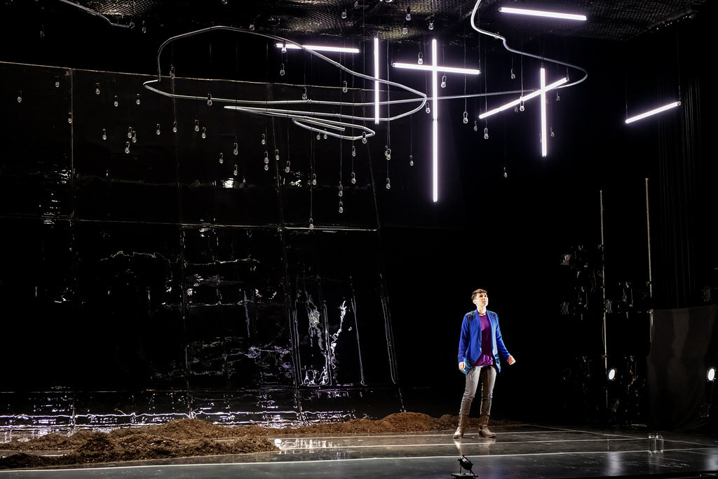 A woman stands in an expansive dark space among small piles of rubble; various tubular fluorescent lights hang above her.