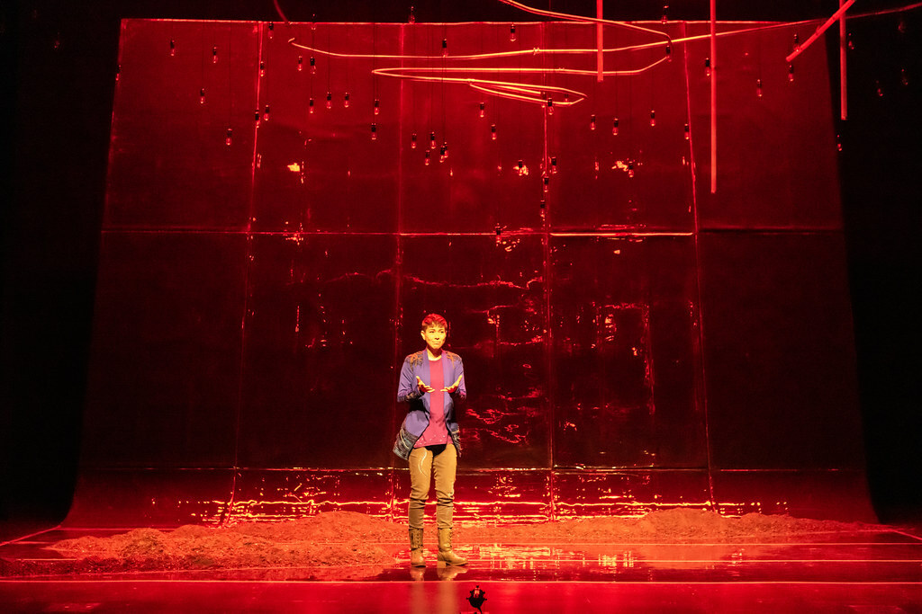 A woman stands before a vast reflective wall bathed in warm amber light, holding her upturned open palms in front of her.