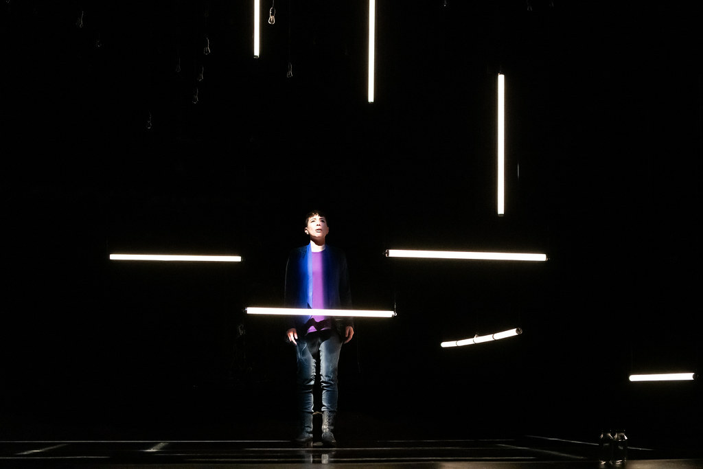 A woman stands half in light and half in shadow; various tubular fluorescent lights surround her.