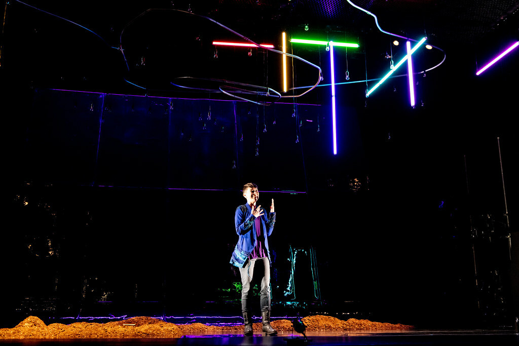 A woman stands on stage among small piles of rubble; colored tubular fluorescent lights hang above her at various angles.