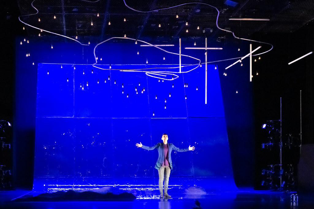 A woman stands before a vast reflective wall bathed in deep blue light, her arms outstretched.