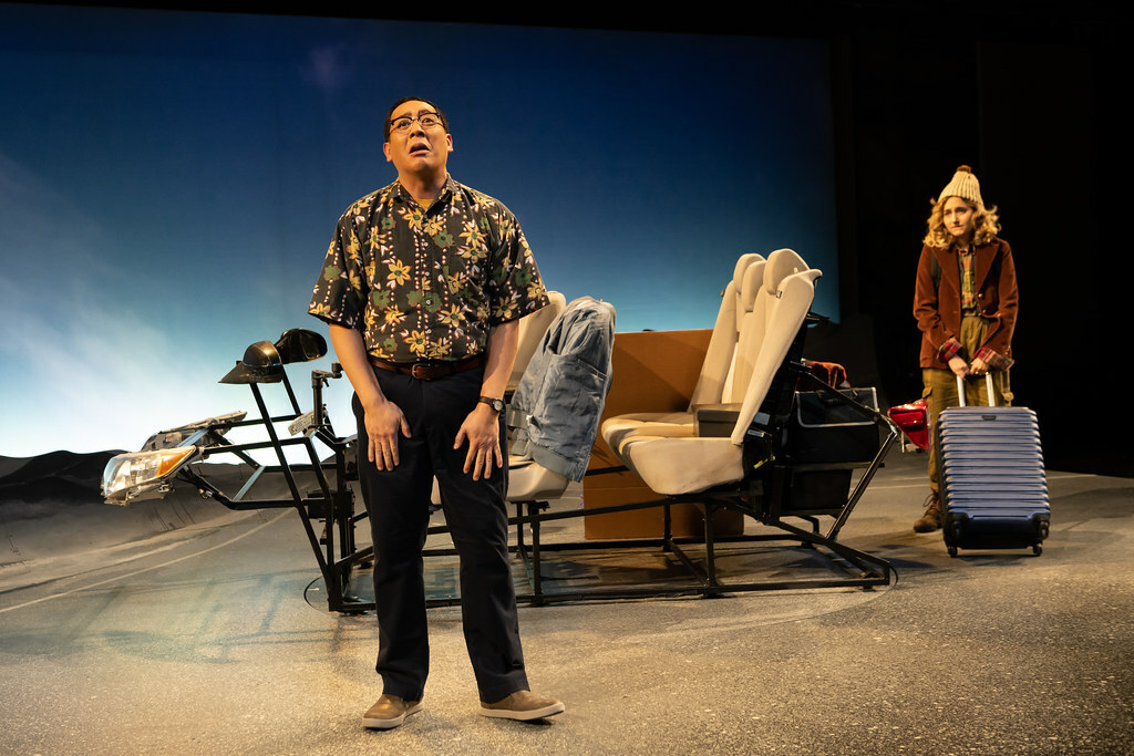 A distraught man in a floral print shirt stands on stage in front of a car, as a woman with a suitcase looks on.