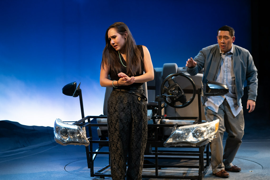 A distraught woman stands on stage in front of a car, as a man looks on from behind, reaching his hand out with concern.