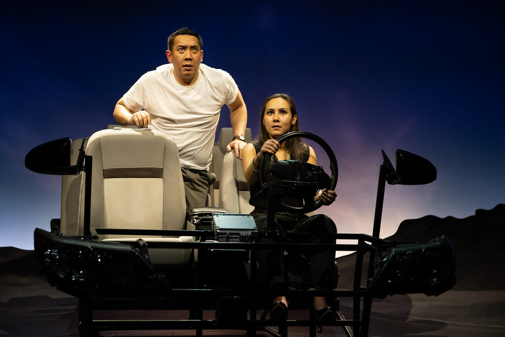 A woman is behind the wheel of a car on stage as a man leans forward from the backseat, both gazing out toward the audience..