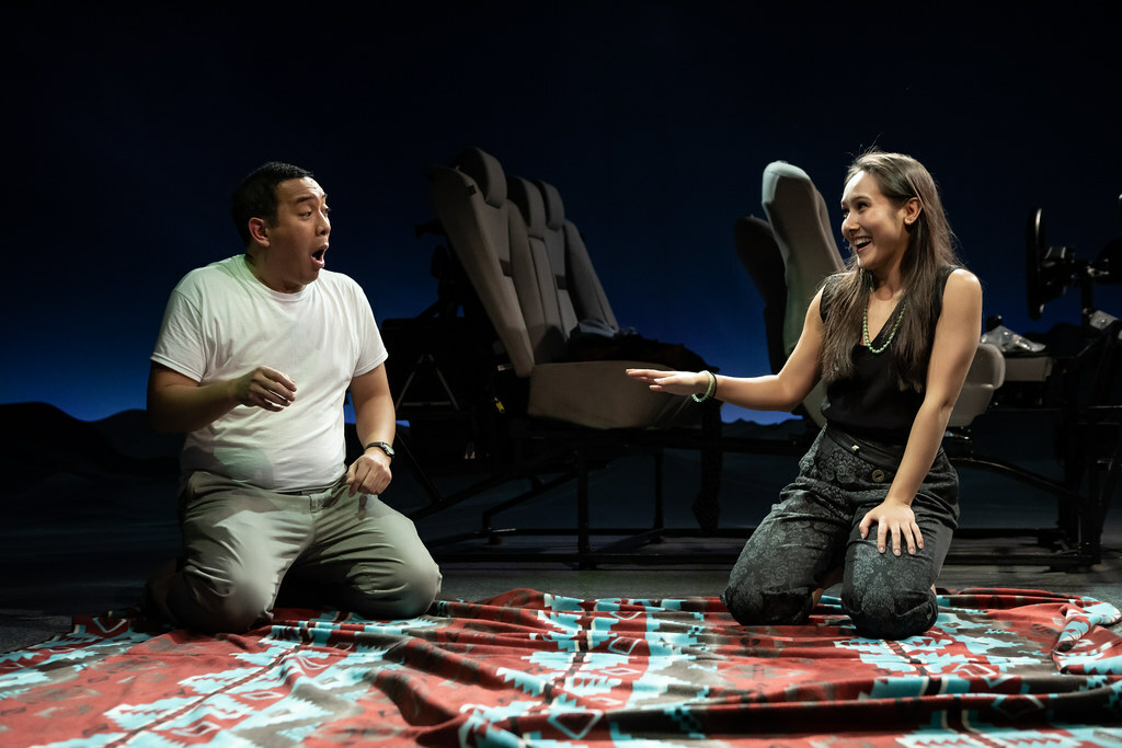 A man and a woman kneel on a colorful blanket spread on the ground. Behind them is a parked car and a dusky horizon beyond.