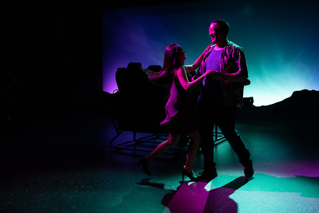 A smiling man and woman dance in the glow of a pink spotlight. Behind them in silhouette are a parked car and dusky horizon.