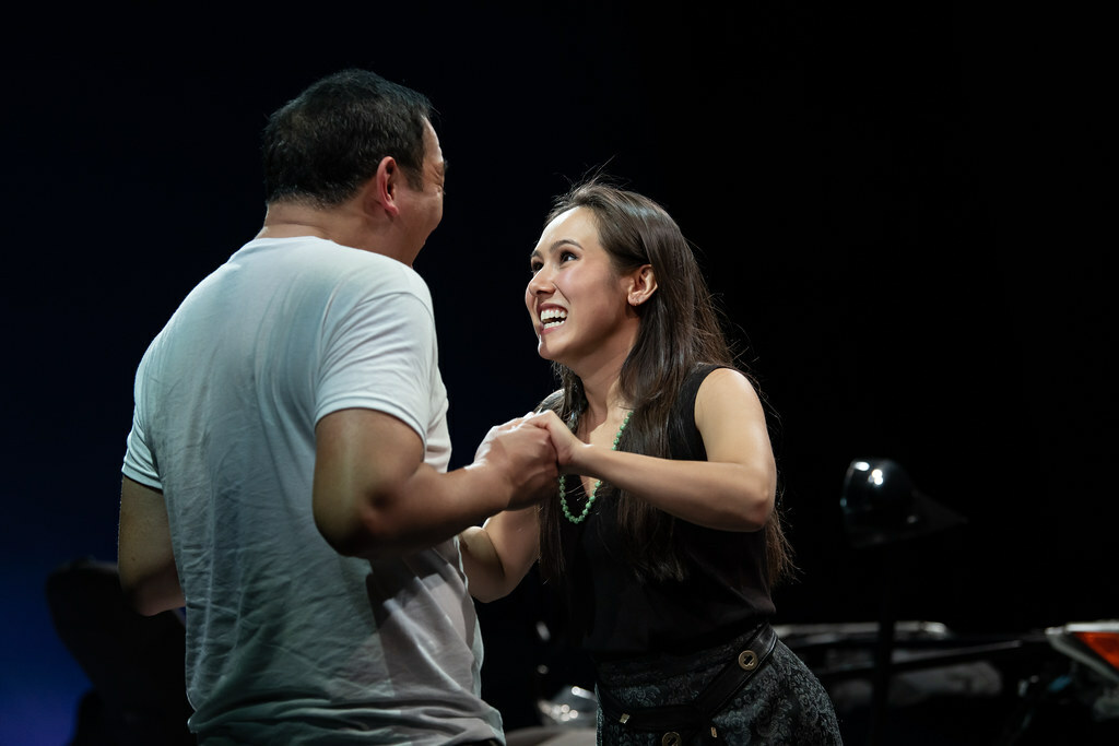 A woman in black and a man in a white t-shirt face each other, smiling excitedly and grasping both of each other's hands.